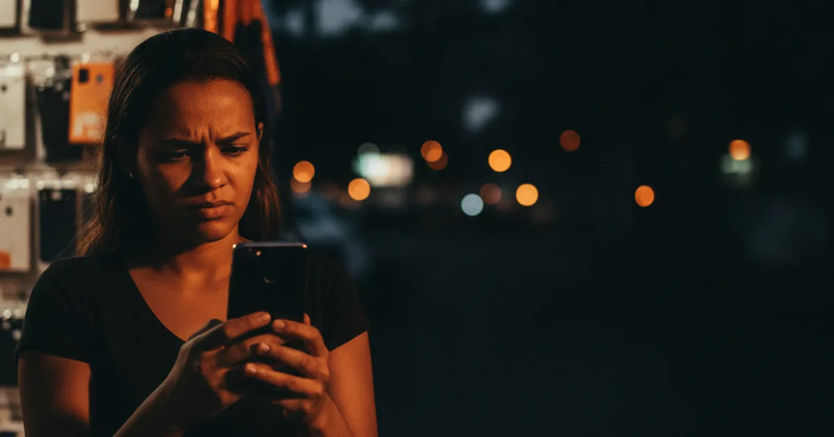 Mulher examinando com desconfiança um celular usado numa banca de rua, com capinhas e acessórios ao fundo